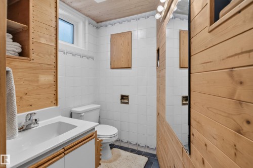 Wood-paneled walls and ceiling, white tiled walls with a decorative border, a white vanity with an integrated sink, and a mirrored wall panel with overhead lighting - 14611 109 Avenue Nw, Edmonton, AB 