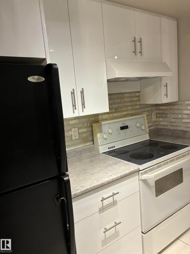 White upper cabinetry with brushed nickel hardware, white range hood, light-toned backsplash, white base cabinetry with brushed nickel hardware, and a black refrigerator - #503 11211 85 St Nw, Edmonton, AB 