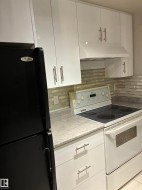 White upper cabinetry with brushed nickel hardware, white range hood, light-toned backsplash, white base cabinetry with brushed nickel hardware, and a black refrigerator - 