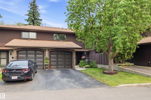 Brown siding townhouse featuring two attached garages with arched brick detailing, a paved driveway, mature trees, and landscaping - 141 Hearthstone Nw, Edmonton, AB - Outdoor