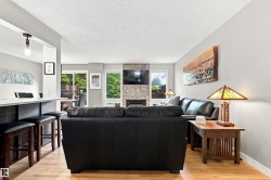 Open-concept living area featuring light wood-finish flooring, a stone-clad fireplace, and multiple windows - 
