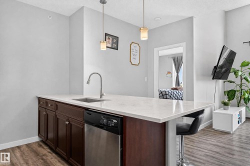 Kitchen island with a light-toned countertop, integrated sink with a gooseneck faucet, dark wood-finish cabinetry, and a stainless steel dishwasher - #411 304 Ambleside Link Li Sw, Edmonton, AB 