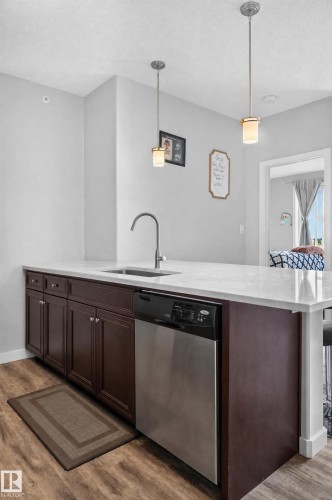 Kitchen island featuring dark wood cabinetry, a stainless steel dishwasher, a single-basin sink with a gooseneck faucet, and white countertops - #411 304 Ambleside Link Li Sw, Edmonton, AB 