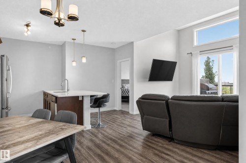 Open-concept living space featuring wood-finish flooring, a kitchen island with seating, stainless steel refrigerator, and large windows with a transom - #411 304 Ambleside Link Li Sw, Edmonton, AB 