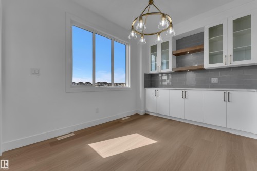 Wood-finish flooring with white baseboards, large multi-pane window, modern brass chandelier, built-in cabinetry with glass-front uppers, and gray subway tile backsplash - 4212 63 Street, Beaumont, AB 