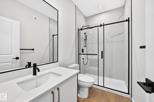 Modern bathroom with a white vanity, integrated sink, matte black fixtures, wood-finish flooring, and a glass-enclosed shower with subway tile surround - 9 Tilia Place, Spruce Grove, AB 
