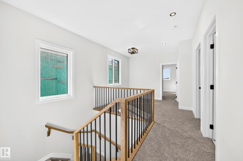 Carpeted hallway featuring a wood and metal baluster staircase, white walls, recessed ceiling lighting, and a decorative flush-mount fixture - 9 Tilia Place, Spruce Grove, AB 