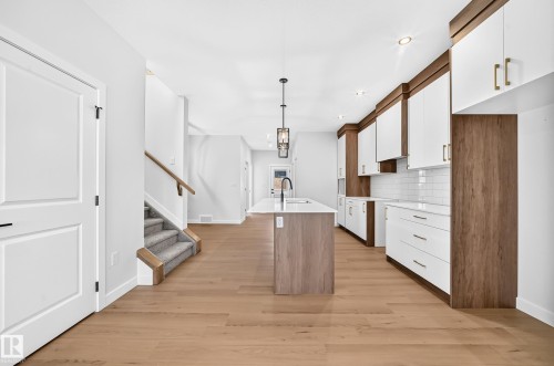 Modern kitchen featuring an island with a built-in sink, two-tone cabinetry with gold-finish hardware, subway tile backsplash, and light wood-finish flooring - 9 Tilia Place, Spruce Grove, AB 