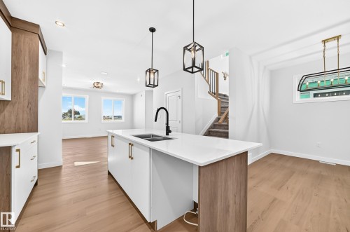 Kitchen island with white countertop and integrated sink, wood-finish flooring, white cabinetry with gold-tone hardware, and a staircase with a wood handrail - 9 Tilia Place, Spruce Grove, AB 