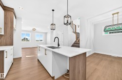 Kitchen island with white countertop and integrated sink, wood-finish flooring, white cabinetry with gold-tone hardware, and a staircase with a wood handrail - 