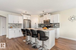 Modern kitchen featuring a large central island with seating, white cabinetry, stainless steel appliances, a mosaic tile backsplash, and wood-finish flooring - 