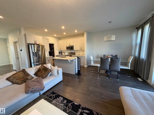 Open-concept living space with dark wood-finish flooring, recessed lighting, and a kitchen featuring white cabinetry and stainless steel appliances - 2628 Donaghey Crescent Sw, Edmonton, AB 