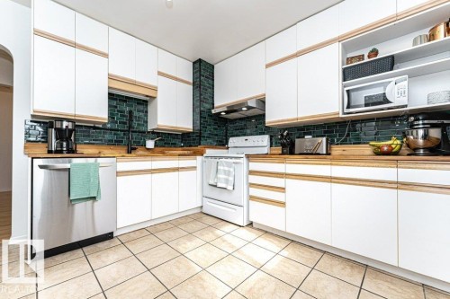 White cabinetry with wood-finish accents, dark subway tile backsplash, wood-finish countertops, stainless steel dishwasher, and neutral tile flooring - 10507 67 Avenue Nw, Edmonton, AB 