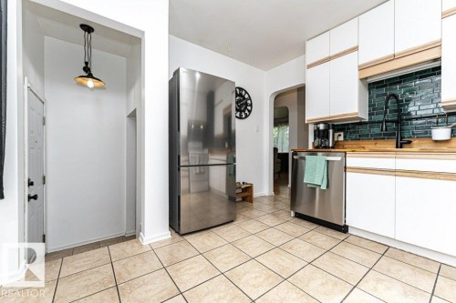 Stainless steel refrigerator and dishwasher, green subway tile backsplash, white cabinetry with wood-finish accents, and tile flooring - 10507 67 Avenue Nw, Edmonton, AB 