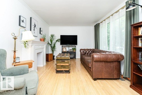 Light wood-finish flooring, white walls, decorative fireplace, crown molding, and large window with sheer and solid drapes - 10507 67 Avenue Nw, Edmonton, AB 