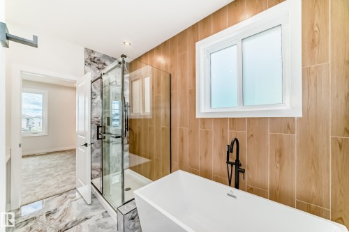 Freestanding soaking tub with matte black floor-mounted faucet, glass-enclosed shower with black fixtures, wood-look tile accent wall, and marble-look floor tiling - 47 Meadowgrove Lane, Spruce Grove, AB 