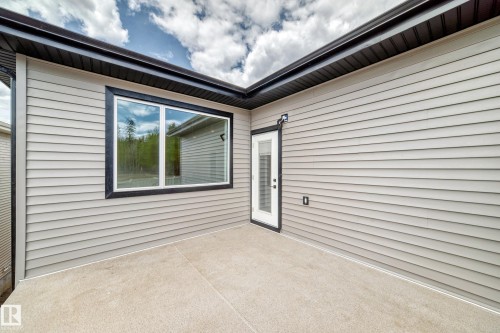Exterior patio featuring concrete surfacing, light-toned horizontal siding, a large fixed window with black trim, and an exterior door with integrated blinds - 47 Meadowgrove Lane, Spruce Grove, AB 