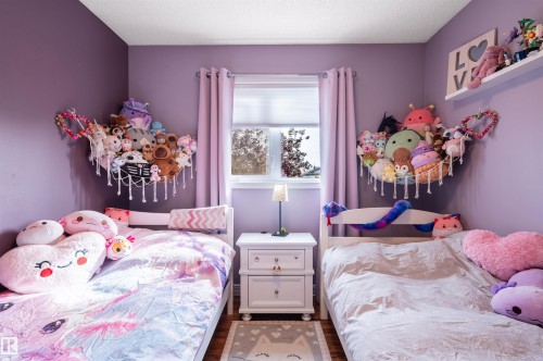 Charming dual bedroom with lavender walls, two beds with coordinating bedding, wood-finish flooring, and a central window with purple curtains providing natural light - 38 Dubonnet Way, St. Albert, AB 