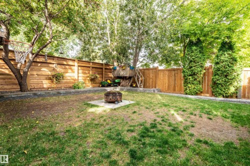 Expansive backyard featuring mature trees, wood privacy fencing, a built-in fire pit area with stone pavers, and concrete retaining walls - 38 Dubonnet Way, St. Albert, AB 