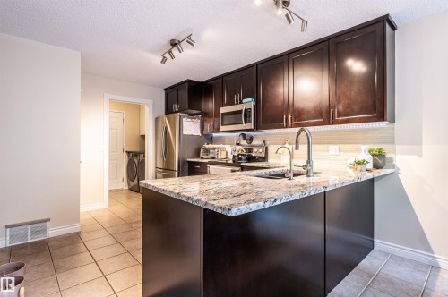 Dark wood-finish cabinetry with stainless steel appliances, speckled stone countertops, and a modern gooseneck faucet - 38 Dubonnet Way, St. Albert, AB 