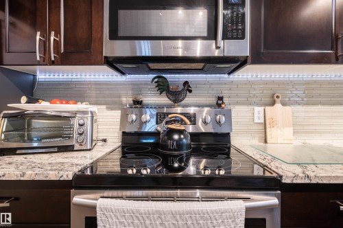 Stainless steel microwave and range, wood-finish cabinetry, stone countertop, and linear tile backsplash - 38 Dubonnet Way, St. Albert, AB 