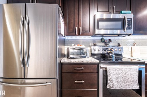 Stainless steel French door refrigerator, dark wood-finish cabinetry, stainless steel microwave, stone-finish countertops, and tile backsplash - 38 Dubonnet Way, St. Albert, AB 