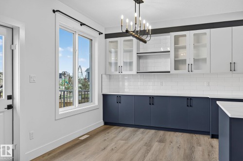 Wood-finish flooring, white subway tile backsplash, two-tone cabinetry with glass inserts, white countertops, and a contemporary chandelier - 22 Sydwyck Circle, Spruce Grove, AB 