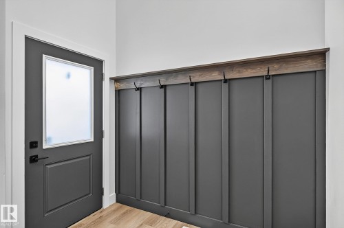 Entryway featuring a dark gray door with a frosted glass panel, matte black hardware, wood-finish flooring, and a built-in board and batten wall with a wood-finish top and integrated hooks - 22 Sydwyck Circle, Spruce Grove, AB 