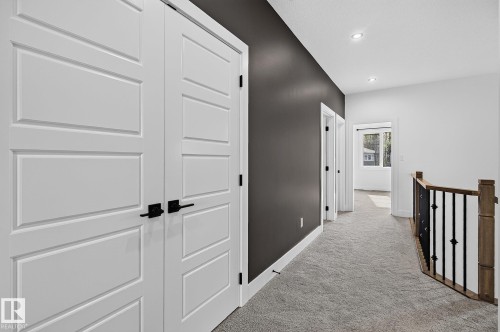 Recessed lighting illuminates the carpeted hallway, featuring a contrasting accent wall, paneled doors with matte black hardware, and a wood-finished railing with black iron balusters - 22 Sydwyck Circle, Spruce Grove, AB 