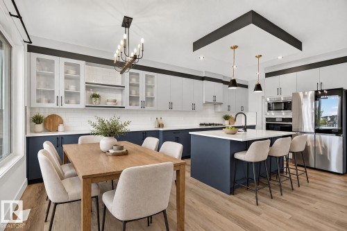 Open-concept kitchen and dining area featuring wood-finish flooring, two-tone cabinetry with glass-front upper cabinets, a kitchen island with seating, and stainless steel appliances - 22 Sydwyck Circle, Spruce Grove, AB 