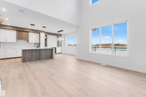 Vaulted ceiling with a large window, light wood-finish flooring, and a kitchen featuring a central island, white and wood-finish cabinetry, and contemporary pendant lighting - 13 Trill Point(E), Spruce Grove, AB 