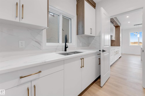 Kitchen featuring white cabinetry with gold-tone hardware, a dual basin sink with a black faucet, white countertops, and a white tile backsplash - 13 Trill Point(E), Spruce Grove, AB 