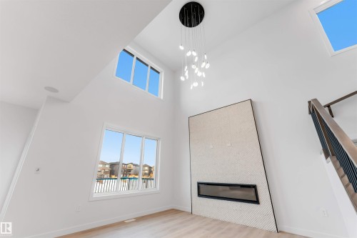 Vaulted ceilings, large windows, a built-in electric fireplace with textured surround, wood-finish flooring, and a modern chandelier - 13 Trill Point(E), Spruce Grove, AB 