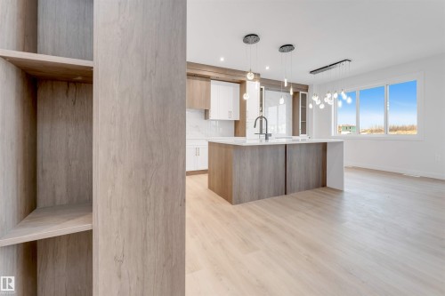 Open-concept living area with light wood-finish flooring, a kitchen island featuring a waterfall countertop and integrated sink, pendant lighting, and abundant natural light from large windows - 13 Trill Point(E), Spruce Grove, AB 