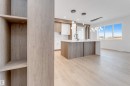 Open-concept living area with light wood-finish flooring, a kitchen island featuring a waterfall countertop and integrated sink, pendant lighting, and abundant natural light from large windows - 13 Trill Point(E), Spruce Grove, AB 