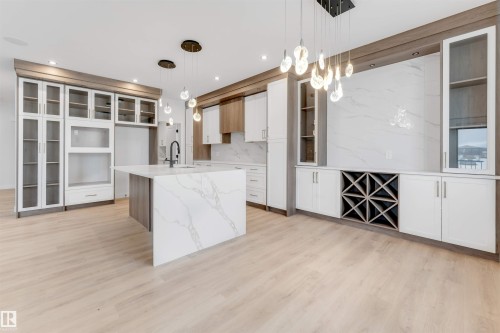 Spacious kitchen featuring a large waterfall island with integrated sink, light wood-finish flooring, two-tone cabinetry, a built-in wine rack, and pendant lighting - 13 Trill Point(E), Spruce Grove, AB 