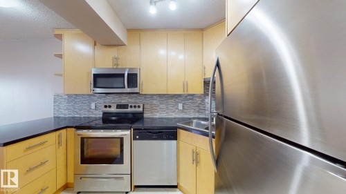 Contemporary kitchen featuring light wood-finish cabinetry, dark countertops, and a mosaic tile backsplash - #307 10140 120 Street Nw, Edmonton, AB - Indoor Photo Showing Kitchen With Upgraded Kitchen