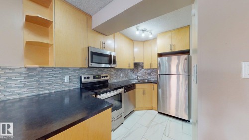 Kitchen featuring light wood cabinetry, dark countertops, a mosaic tile backsplash, and stainless steel appliances - #307 10140 120 Street Nw, Edmonton, AB - Indoor Photo Showing Kitchen