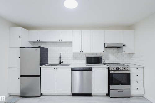 White shaker cabinetry with dark hardware, stainless steel appliances, dual basin sink with matte black faucet, white subway tile backsplash, and light-toned flooring - 11134 11138 116 Street Nw, Edmonton, AB 