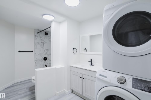 White stacked laundry appliances, vanity with a white countertop, wall-mounted mirror, and a shower with patterned wall tile - 11134 11138 116 Street Nw, Edmonton, AB 