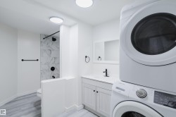 White stacked laundry appliances, vanity with a white countertop, wall-mounted mirror, and a shower with patterned wall tile - 