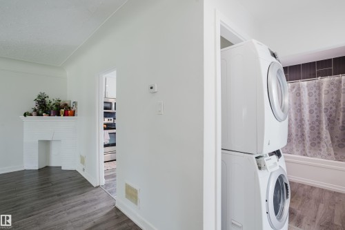 White brick fireplace, wood-finish flooring, white walls, stacked laundry appliances, and a full bathtub with dark tile surround - 11134 11138 116 Street Nw, Edmonton, AB 