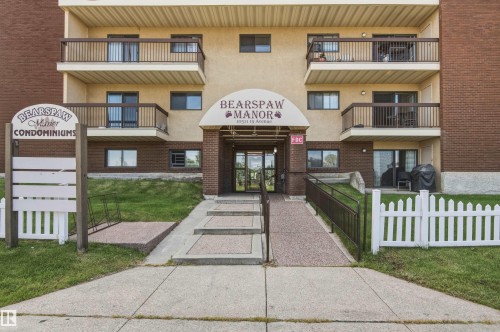 Building exterior with a brick and stucco facade, multiple private balconies, a ground-level patio, a covered main entrance, and a white picket fence - #115 10511 19 Av Nw, Edmonton, AB 