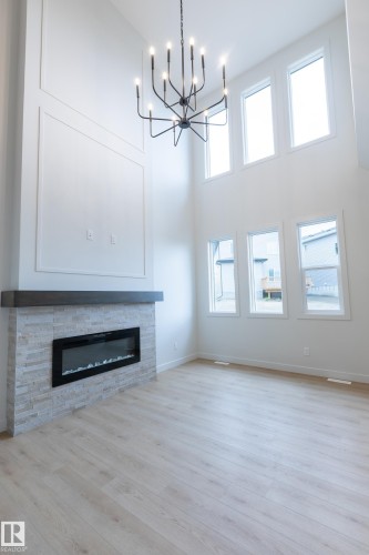Two-story living area featuring a linear electric fireplace with a stacked stone facade and wood mantel, light wood-finish flooring, and a contemporary chandelier - 4005 36 Street, Beaumont, AB 