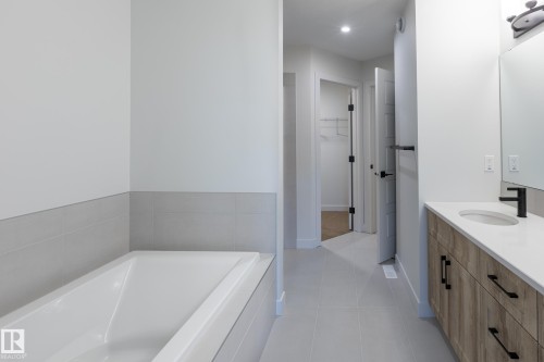 White soaking tub with tiled surround, light gray tile flooring, wood-finish vanity with countertop, rectangular undermount sink, and matte black faucet - 4005 36 Street, Beaumont, AB 