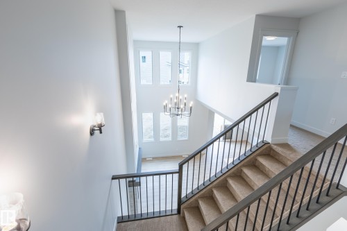 Two-story interior with a carpeted staircase featuring dark metal railings, a suspended chandelier, multiple tall windows, and a wall-mounted sconce - 4005 36 Street, Beaumont, AB 