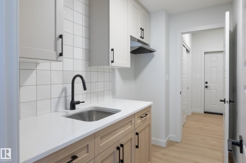 Compact kitchen area featuring a stainless steel sink with a matte black faucet, white square tile backsplash, light-toned wood-finish lower cabinetry, white upper cabinetry, and light wood-finish flooring - 4005 36 Street, Beaumont, AB 