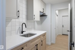 Compact kitchen area featuring a stainless steel sink with a matte black faucet, white square tile backsplash, light-toned wood-finish lower cabinetry, white upper cabinetry, and light wood-finish flooring - 