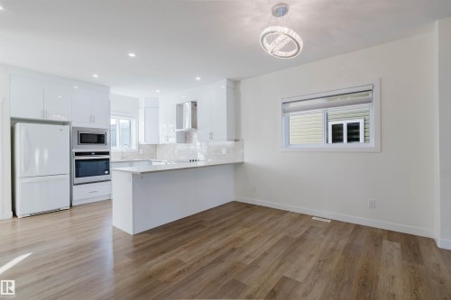 Open-concept kitchen featuring white cabinetry, light countertops, wood-finish flooring, built-in stainless steel appliances, and a modern ceiling light fixture - 6047 180 Avenue Nw, Edmonton, AB 