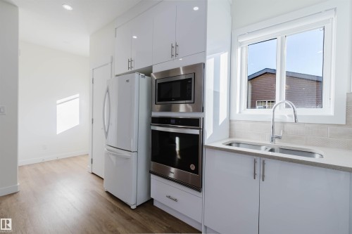 White high-gloss cabinetry, stainless steel built-in appliances, undermount double-basin sink, light-toned countertop, and wood-finish flooring - 6047 180 Avenue Nw, Edmonton, AB 